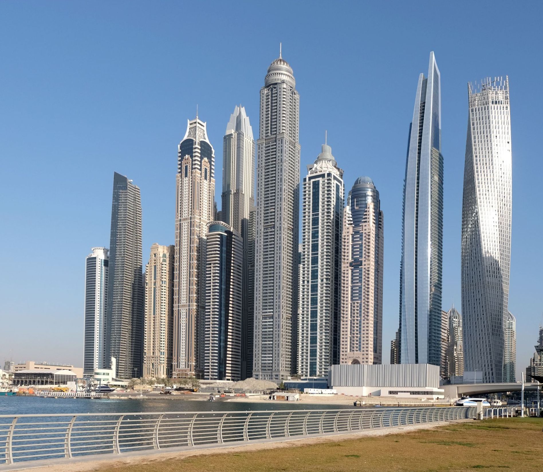 Aedas’s DAMAC Heights in Dubai is shown as a tall residential skyscraper with a vertically emphasised profile, continuous glass curtain wall, and a distinctive stepped crown. The exterior highlights high‑rise residential architecture integrated into Dubai Marina’s dense skyline, with articulated balcony bands and a tapered upper massing.