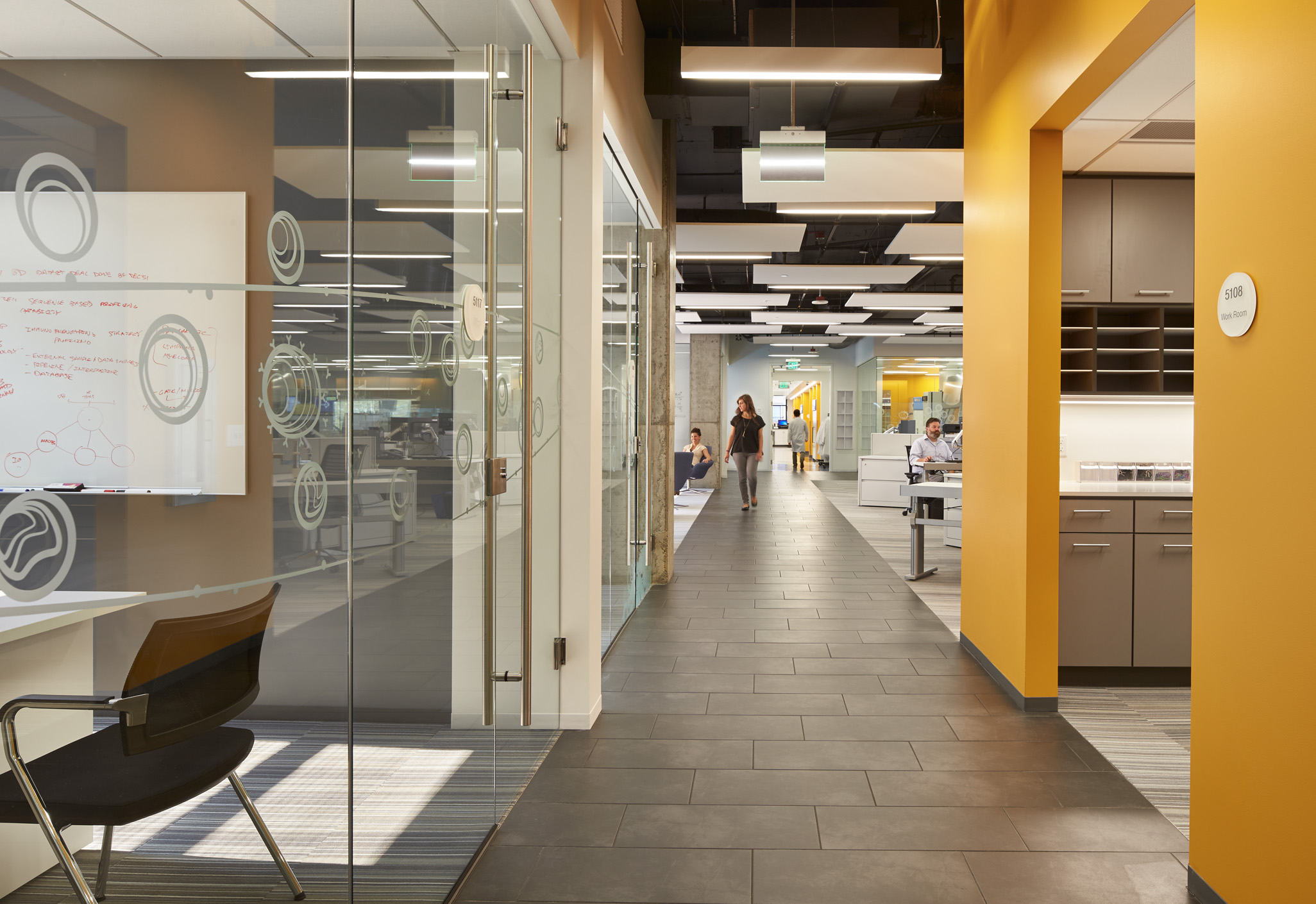 Aedas’s Celgene Seattle workplace interior is shown as an open‑plan office environment with glazed conference rooms, linear circulation spine, exposed services ceiling, and integrated workstations. The image highlights flexible research‑support architecture, transparent partition systems, and performance‑led workplace planning within a life‑sciences headquarters.