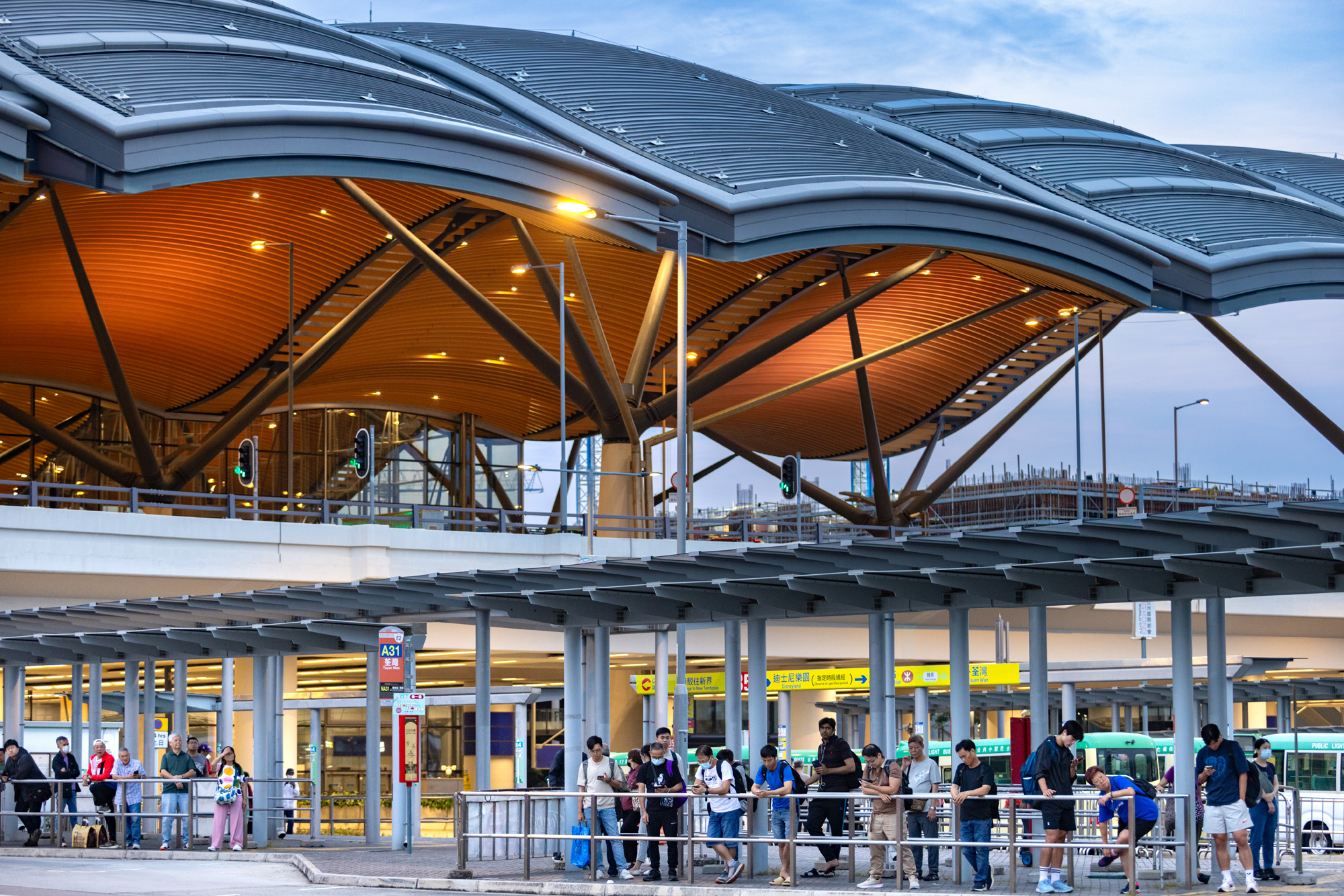 Aedas’ HZMB Hong Kong Port Passenger Clearance Building with modular wave roof and glazed canyons on reclaimed island. Exterior view of the HZMB Hong Kong Port Passenger Clearance Building designed by Aedas, featuring a modular wave‑form roof supported by steel columns. Full‑height glazed ends and internal canyons allow daylight penetration. The building sits on a reclaimed island, with covered forecourts, transport lanes, and wayfinding elements supporting high‑volume border‑crossing operations.