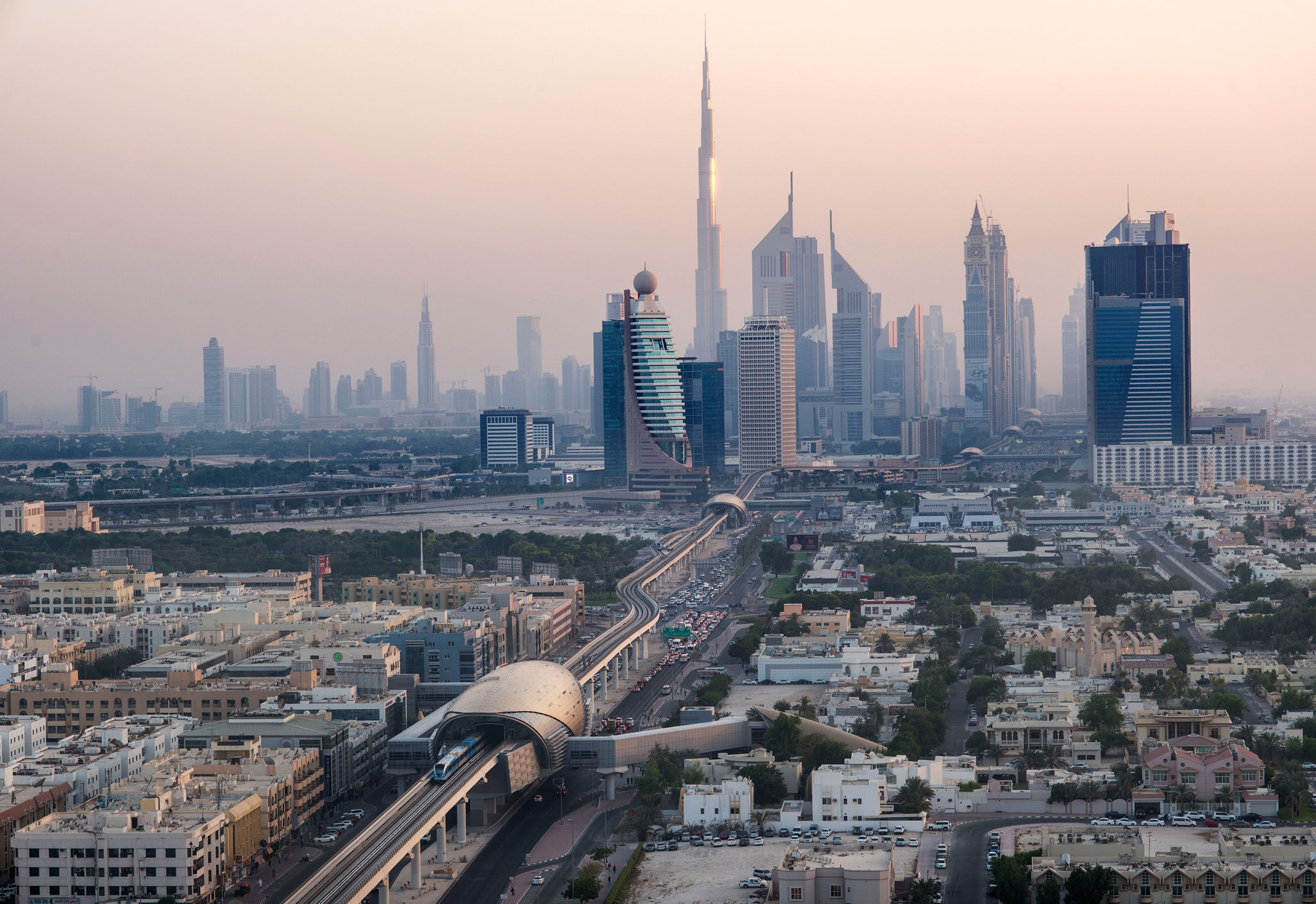 Aedas’ elevated Dubai Metro Red Line station with pearl-shell canopy enclosing platforms along Sheikh Zayed Road, Dubai. Aerial view of an Aedas-designed Dubai Metro elevated station, showing an armadillo-like metallic shell canopy spanning dual rail tracks. The station sits above a multi-lane arterial road, forming a linear transit spine through dense urban fabric, with prefabricated structural ribs enclosing platforms and concourse levels in a hot-arid desert climate.