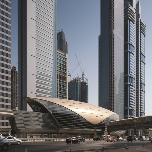 Aedas’ elevated Dubai Metro Red Line station with pearl-shell canopy enclosing platforms along Sheikh Zayed Road, Dubai. Aerial view of an Aedas-designed Dubai Metro elevated station, showing an armadillo-like metallic shell canopy spanning dual rail tracks. The station sits above a multi-lane arterial road, forming a linear transit spine through dense urban fabric, with prefabricated structural ribs enclosing platforms and concourse levels in a hot-arid desert climate.