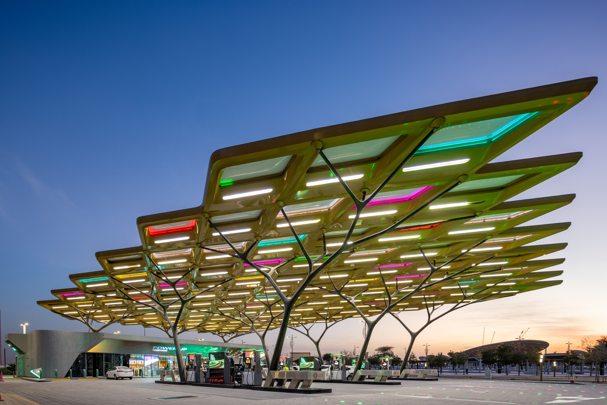 Aedas’ ENOC Future Station – Dubai Expo 2020, Dubai: Ghaf-inspired canopy shading fuel bays on a wide-span, tree-column structure. Twilight view of Aedas-designed ENOC Future Mobility Station forecourt in Dubai, with a Ghaf-tree canopy expressed as a diamond-grid soffit on branching columns. Open fuel islands sit beneath integrated lighting; a low retail volume anchors the edge, and paved circulation zones frame vehicle approach at a wide-span, shade-optimised canopy footprint.