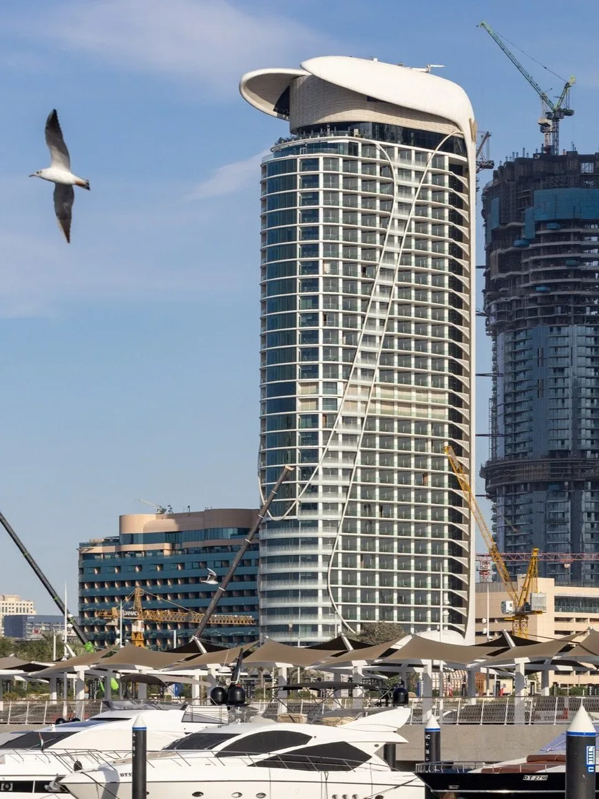 Aedas’ W Dubai – Mina Seyahi beachfront hotel tower with curved glazed façade overlooking Dubai Marina. Harbour‑edge view of the Aedas‑designed W Dubai – Mina Seyahi showing a 31‑storey hotel tower with a curved profile and high‑performance glazed façade. The building stands adjacent to marina berths and yachts, with a dedicated podium and independent drop‑off, set within Dubai’s coastal, high‑rise waterfront skyline and arid maritime climate.
