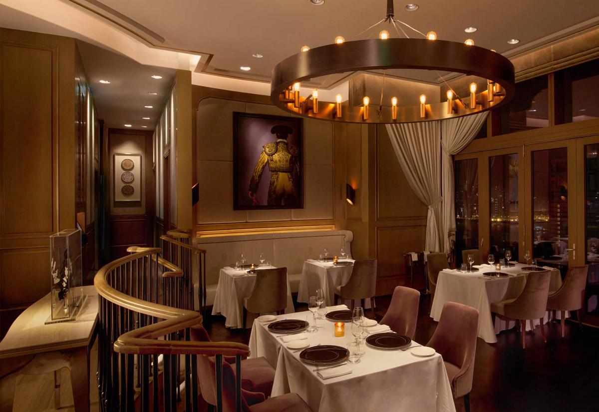 Aedas Interiors’ Zurriola in Hong Kong is shown as a contemporary fine‑dining interior with soft fabric‑upholstered seating, timber wall panelling, sheer drapery, and a circular chandelier feature. The image highlights an elegant, romantic hospitality environment defined by muted tones, refined lighting, and luxurious textures that create an intimate and serene dining atmosphere.