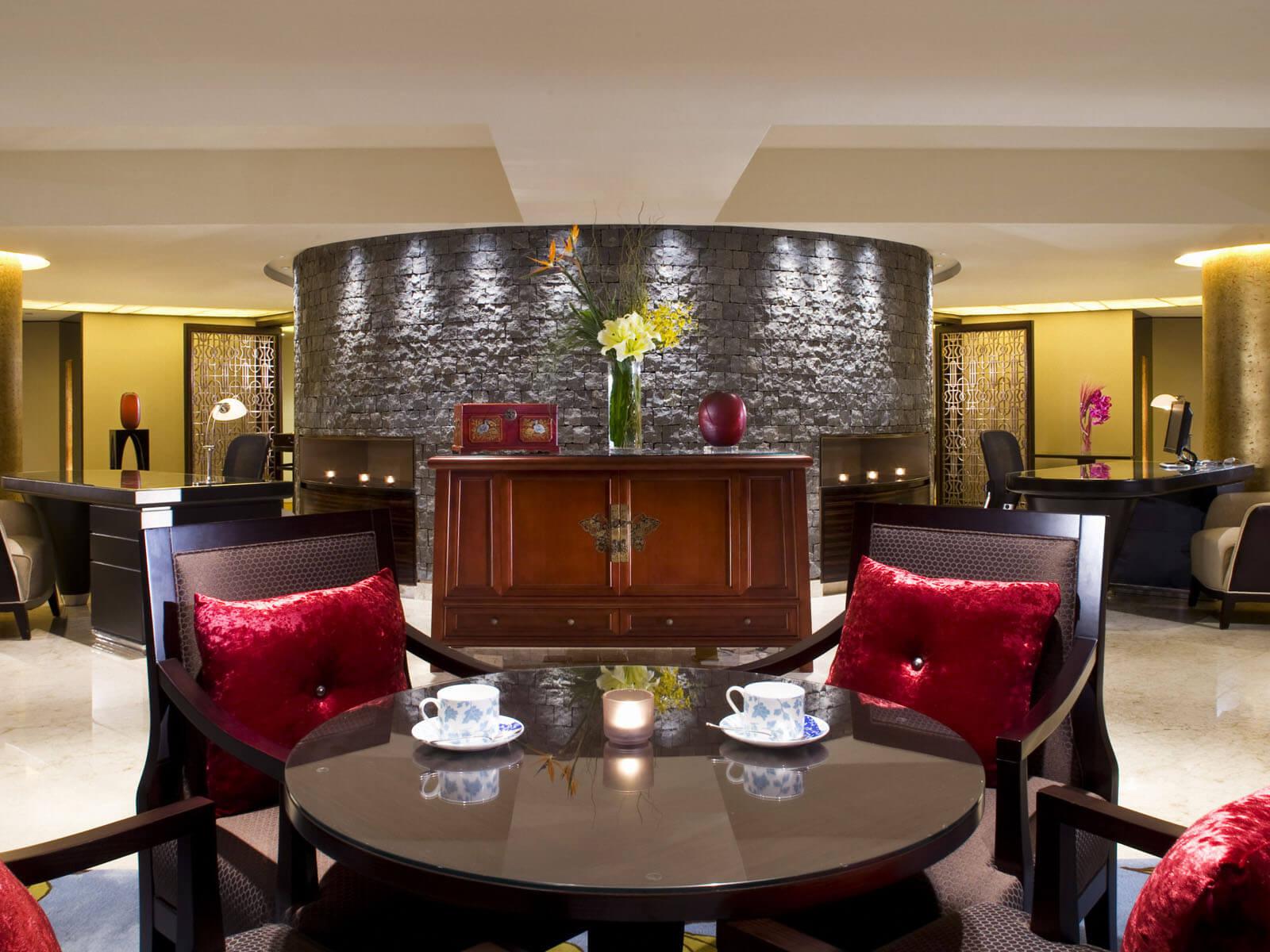 Aedas Interiors’ The Portman Ritz‑Carlton Hotel Shanghai is shown as an intimate lounge setting with curved stone feature walls, dark timber furniture, plush upholstered seating, and warm ambient lighting. The image highlights a refined hospitality interior blending Eastern and Western influences through layered materials, controlled symmetry, and elegant detailing designed to support comfort and privacy within a luxury hotel environment.