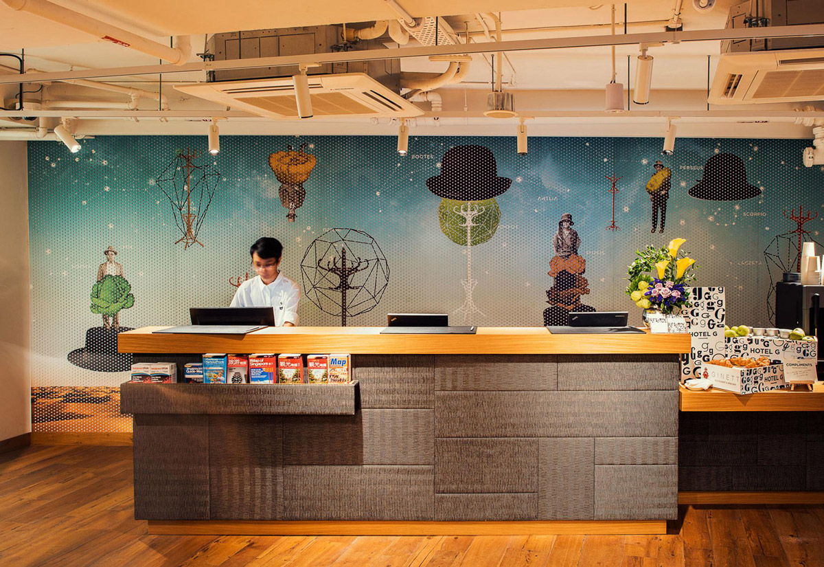 Aedas Interiors’ Hotel G in Singapore is shown with an urban, informal reception and social space featuring an exposed industrial ceiling, long timber reception counter, patterned graphic feature wall, and warm pendant lighting. The image highlights a street‑engaging hospitality interior designed for mingling, casual work, and social interaction, blending raw finishes with playful local references and bespoke furniture.