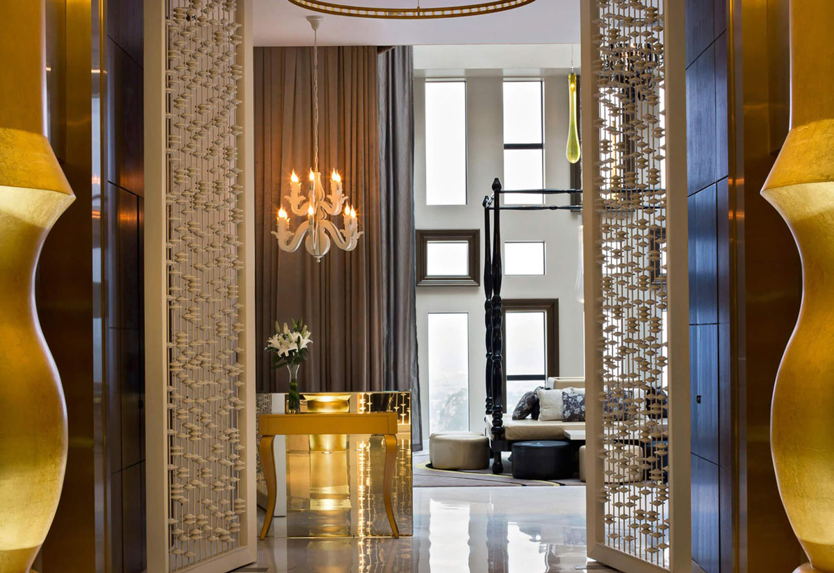 Aedas Interiors’ Scoozi & Twenty‑Six in Liuzhou is shown as a dramatic restaurant arrival with patterned metallic screens, sculptural floor lamps, polished stone flooring, and a crystal chandelier framing a lounge beyond. The image highlights a theatrical hospitality interior featuring layered textures, curated lighting, and a transition from dimmed entry to a luminous double‑height social space with panoramic city views.