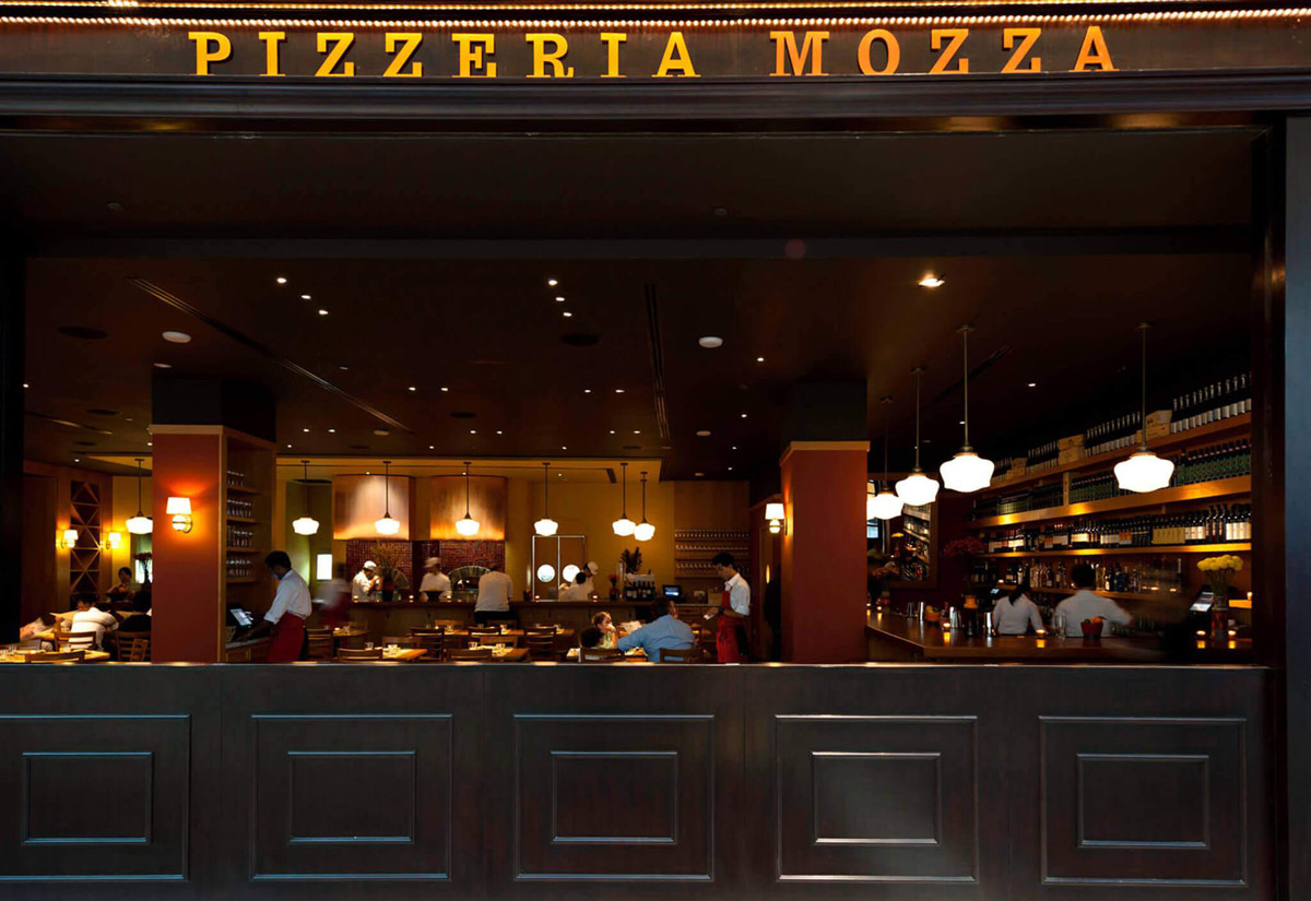 Aedas Interiors’ Pizzeria Mozza in Singapore is shown as a lively, open‑front restaurant interior with a prominent illuminated “PIZZERIA MOZZA” sign, dark timber bar counter, central white marble mozzarella bar, and warm pendant lighting. The image highlights an energetic, urban dining environment where open kitchens, wine displays, and communal seating create a vibrant hospitality experience rooted in Italian culinary tradition.