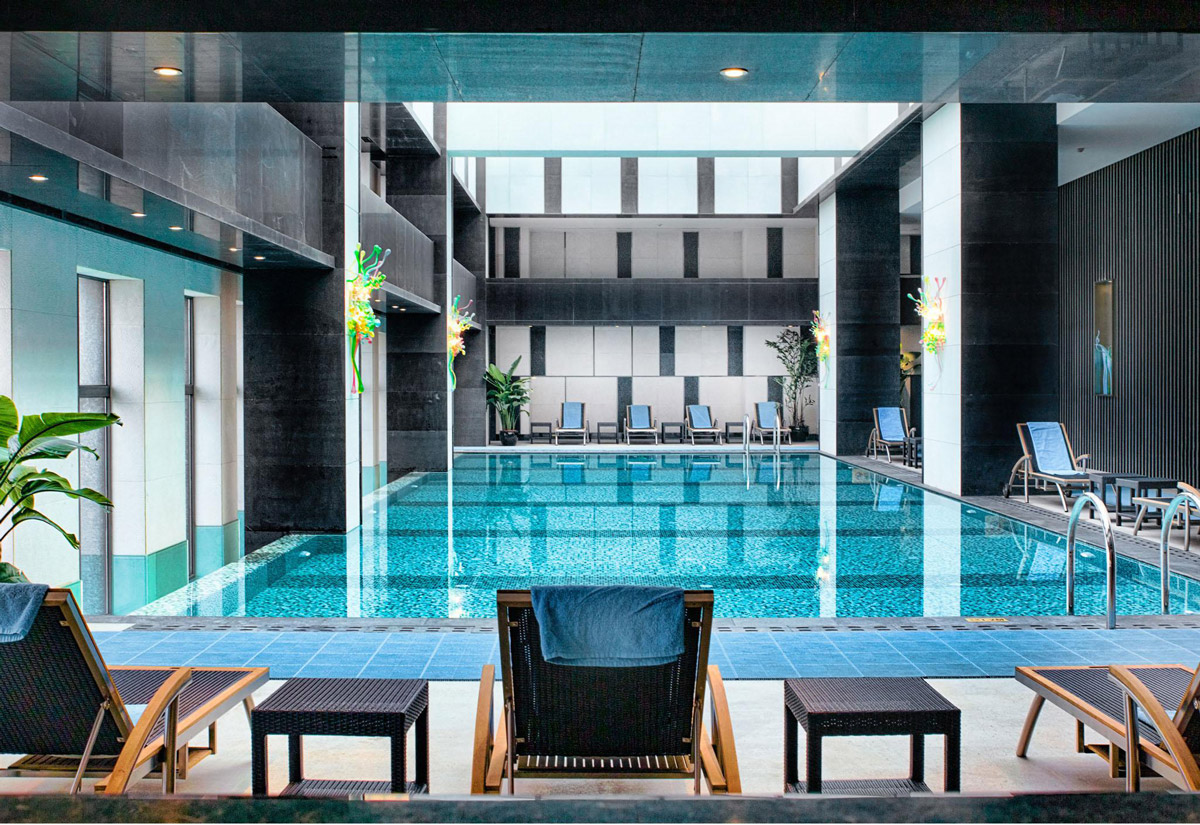 Aedas Interiors’ Ximei Business Hotel in Shijiazhuang is shown with a tranquil indoor rooftop infinity pool featuring gradient mosaic tiling, dark stone‑clad columns, and lounge chairs arranged along the pool edge. The image highlights a contemporary hospitality leisure space combining reflective water surfaces, clean architectural lines, and controlled natural light to create a refined business‑hotel retreat overlooking the urban skyline.