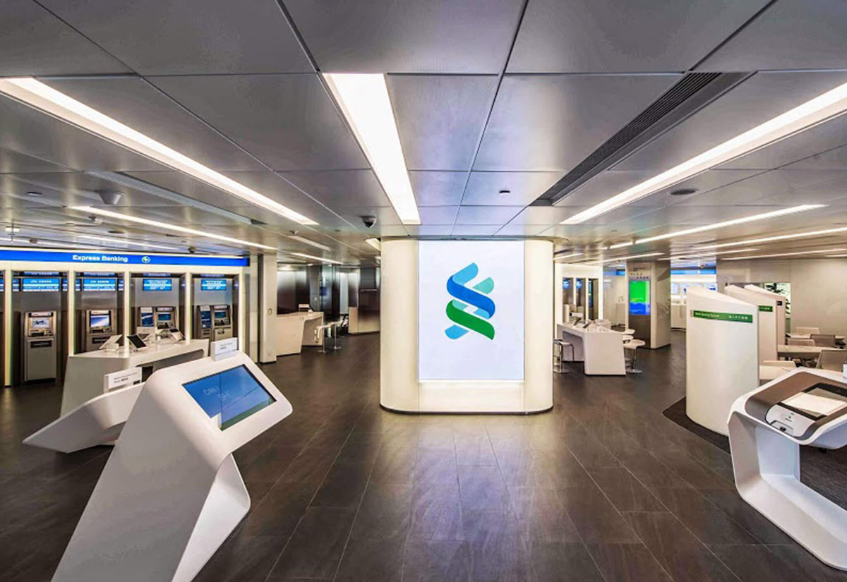 Aedas Interiors’ Standard Chartered Bank Des Voeux Road retail branch in Hong Kong is shown as a modern banking hall with self‑service kiosks, integrated digital ATMs, and a central illuminated branding column. The image highlights a secure, streamlined financial interior featuring neutral finishes, clear circulation, and technology‑enabled customer zones designed to enhance efficiency and brand visibility within a high‑traffic urban branch environment.
