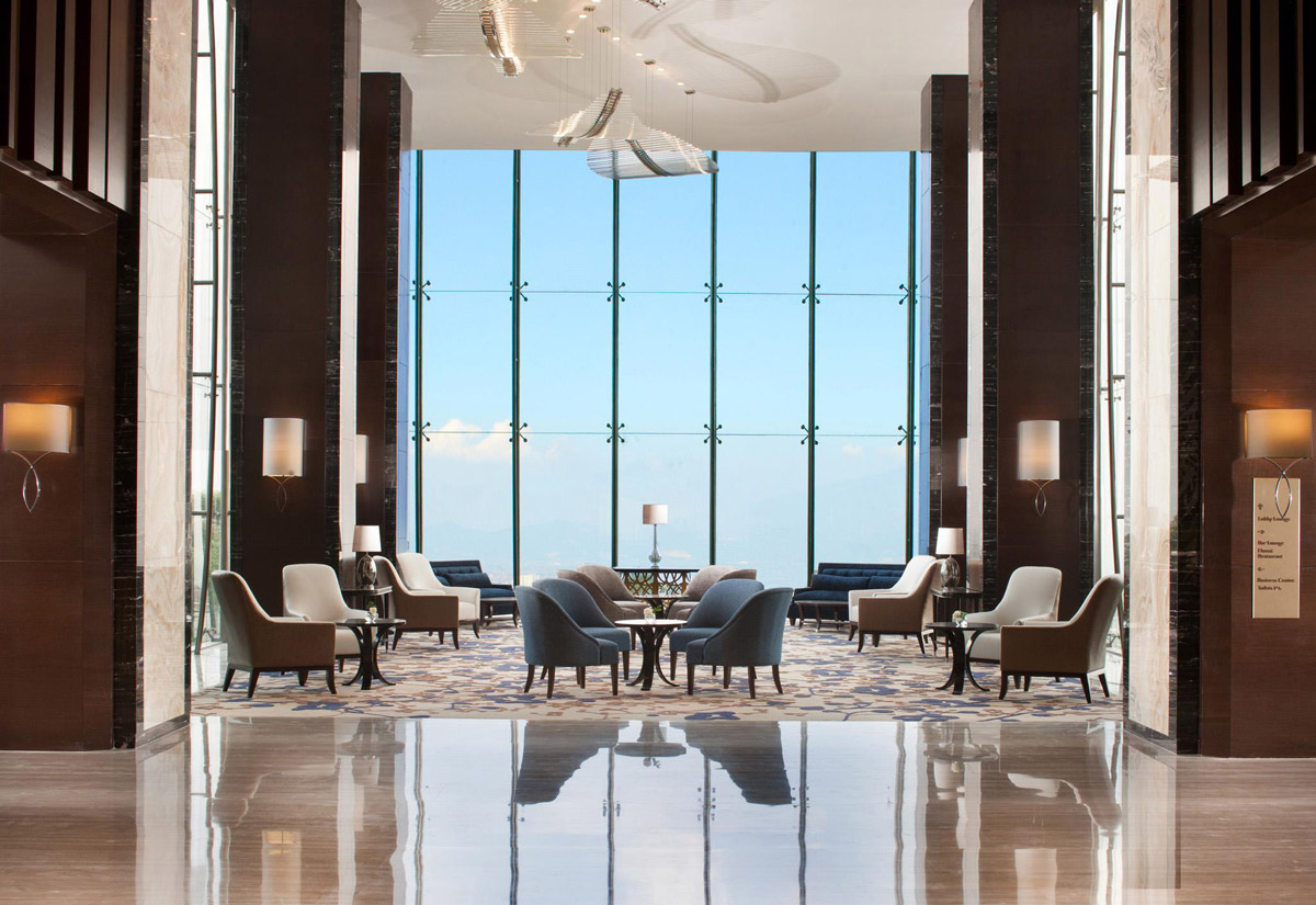 Aedas Interiors’ InterContinental Hotel Bandung is shown as a grand hotel lobby with floor‑to‑ceiling glazing framing natural views, polished stone flooring, dark timber wall panels, and sculptural ceiling art. The image highlights a serene hospitality interior that balances contemporary luxury with a restrained material palette, allowing landscape and light to define the spatial experience.