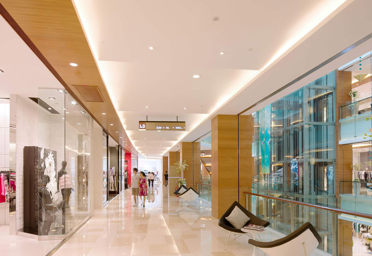 Aedas Interiors’ Renhe Spring Department Store in Chengdu is shown as a refined retail gallery with polished stone flooring, warm timber ceiling panels, full‑height glass shopfronts, and integrated lounge seating overlooking a glazed atrium. The image highlights an upscale shopping environment where natural materials, soft lighting, and generous circulation create a cosmopolitan, welcoming department‑store experience supporting international and independent brands.