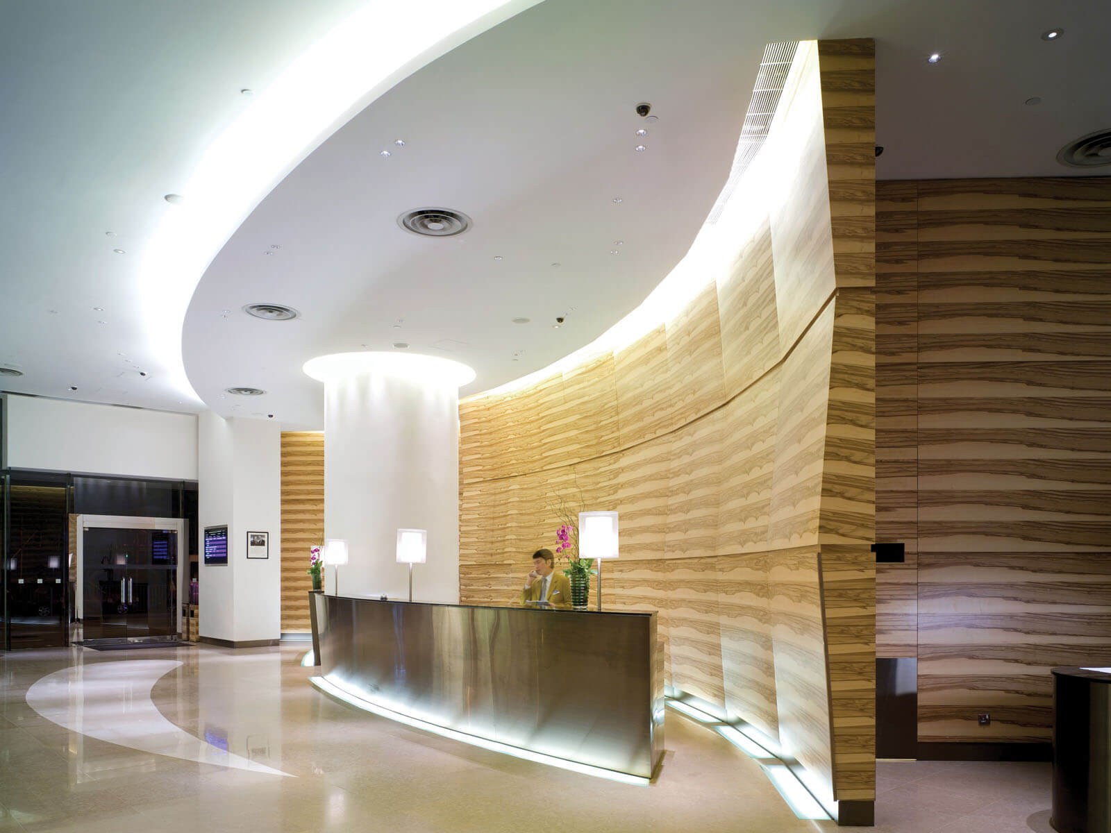 Aedas Interiors’ Novotel Century Hong Kong lobby is shown as a fluid, contemporary arrival space with a curved metallic reception counter, layered stone‑textured wall panels, and sculptural ceiling lighting. The image highlights a water‑inspired hospitality interior where soft curves, reflective surfaces, and warm illumination create a calm yet dynamic focal point within the hotel’s public areas.