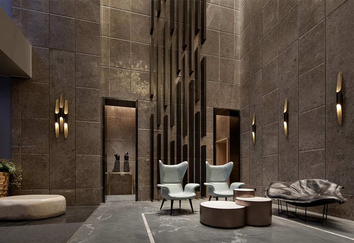 Aedas Interiors’ One Avenue Tianyuan show flat in Shenzhen is shown as a dramatic double‑height interior with stone‑clad walls, sculptural vertical metal installation, refined lounge seating, and soft accent lighting. The image highlights an understated luxury residential environment where European contemporary elegance is blended with Chinese influences through rich materials, geometric spatial order, and bespoke detailing.