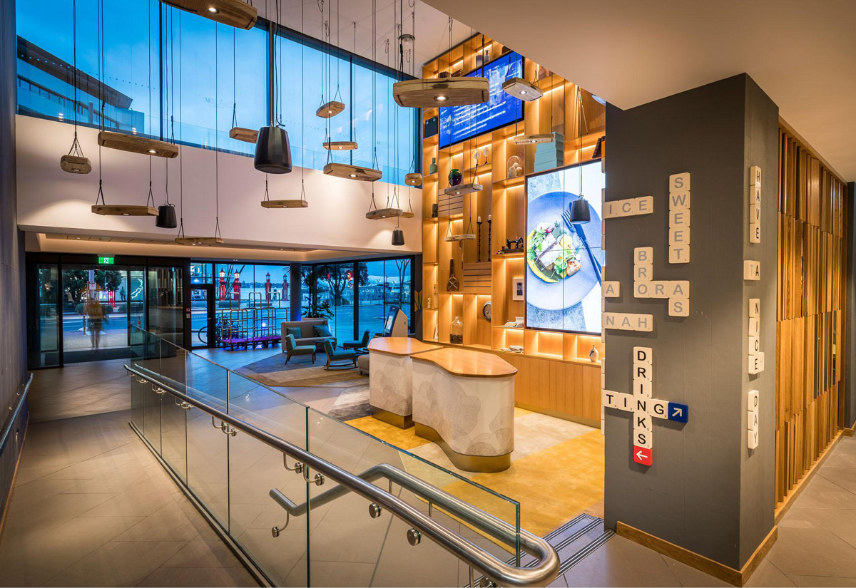 Aedas Interiors’ M Social in Auckland is shown as a vibrant double‑height hotel lobby and dining space with layered timber shelving, illuminated display walls, and sculptural pendant lighting. The image highlights a socially driven hospitality interior oriented toward waterfront views, blending contemporary design with nostalgic local references and open, interactive public spaces that encourage movement, gathering, and casual dining.
