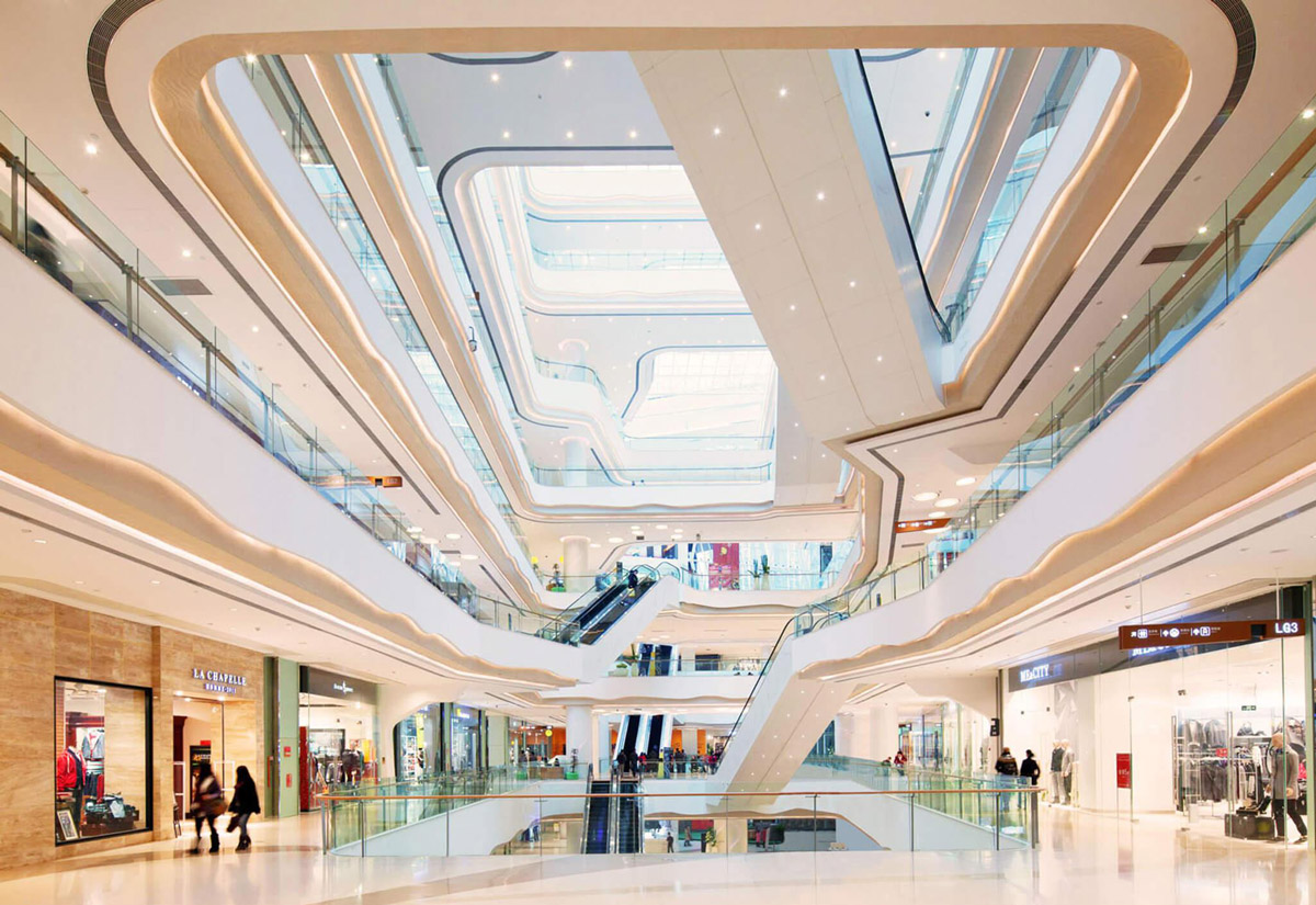 Aedas Interiors’ Starlight Place in Chongqing is shown as a multi‑storey retail atrium with fluid, sculptural white balcony forms, integrated escalators, and continuous ribbon‑like ceiling lighting. The image highlights a dynamic shopping‑mall interior where contrasting curves and straight lines, light and shadow, and smooth surfaces create a visually layered circulation space that draws visitors upward through the mall.