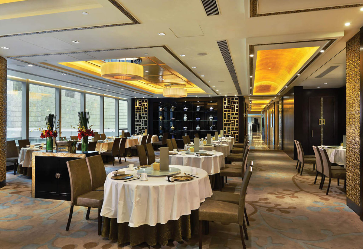 Aedas Interiors’ Ming Court at Langham Place, Hong Kong is shown as an upscale Chinese fine‑dining interior with round banquet tables, upholstered chairs, and a refined palette of warm neutrals. The image highlights silver‑leaf accented wall panels, abstract floor patterns inspired by Chinese characters, illuminated ceiling coffers, and a carefully balanced blend of traditional motifs with contemporary luxury finishes.