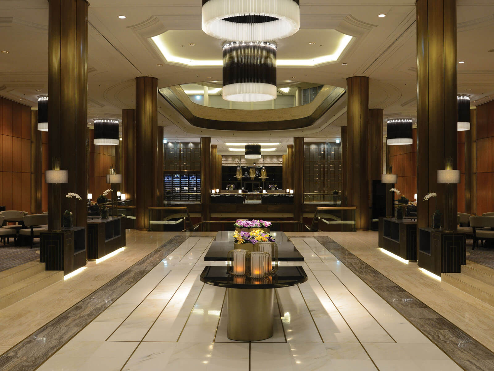 Aedas Interiors’ Millennium Seoul Hilton lobby is shown as a grand, symmetrical hospitality interior with polished marble flooring, tall timber‑clad columns, and layered circular chandeliers. The image highlights a dignified arrival space where original material richness is retained and enhanced through contemporary lighting, refined furniture, and subtle textural updates to refresh the hotel’s timeless architectural identity.