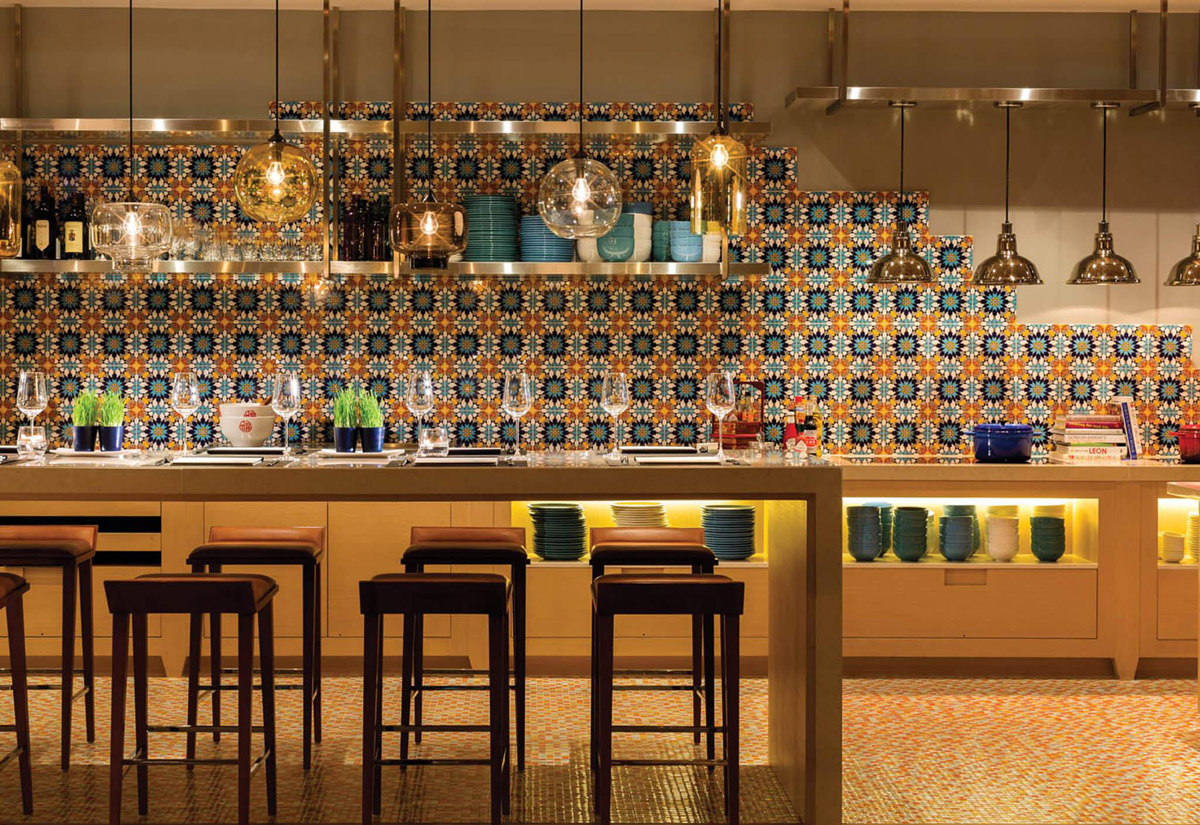 Aedas Interiors’ Café Post at Hotel Indigo Hong Kong Island is shown as a casual all‑day dining interior with an open kitchen bar, patterned ceramic tile backsplash, exposed shelving of tableware, and warm pendant lighting. The image highlights a welcoming, home‑like restaurant environment designed for social interaction, blending contemporary finishes with references to local heritage in a relaxed hospitality setting.