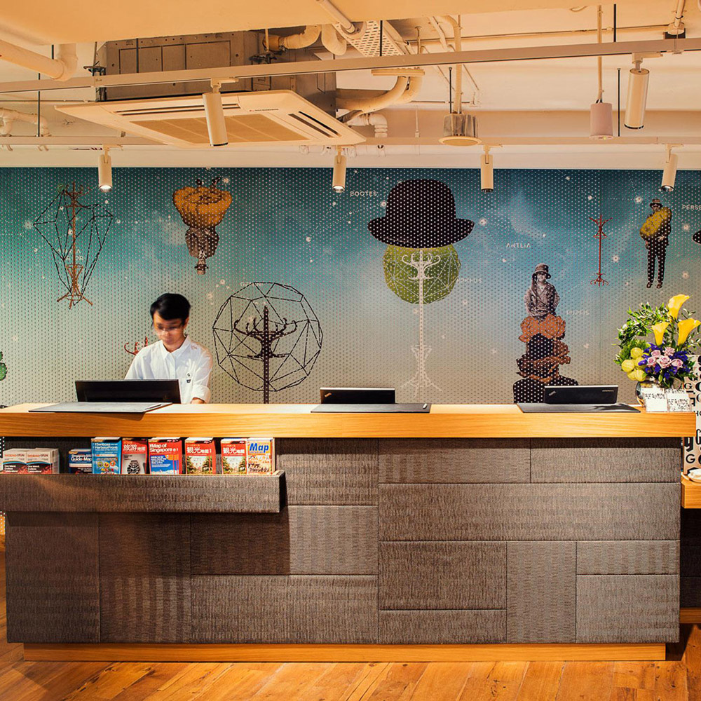 Aedas Interiors’ Hotel G in Singapore is shown with an urban, informal reception and social space featuring an exposed industrial ceiling, long timber reception counter, patterned graphic feature wall, and warm pendant lighting. The image highlights a street‑engaging hospitality interior designed for mingling, casual work, and social interaction, blending raw finishes with playful local references and bespoke furniture.