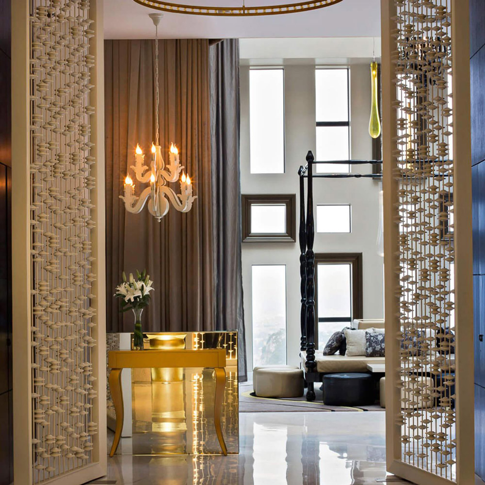 Aedas Interiors’ Scoozi & Twenty‑Six in Liuzhou is shown as a dramatic restaurant arrival with patterned metallic screens, sculptural floor lamps, polished stone flooring, and a crystal chandelier framing a lounge beyond. The image highlights a theatrical hospitality interior featuring layered textures, curated lighting, and a transition from dimmed entry to a luminous double‑height social space with panoramic city views.