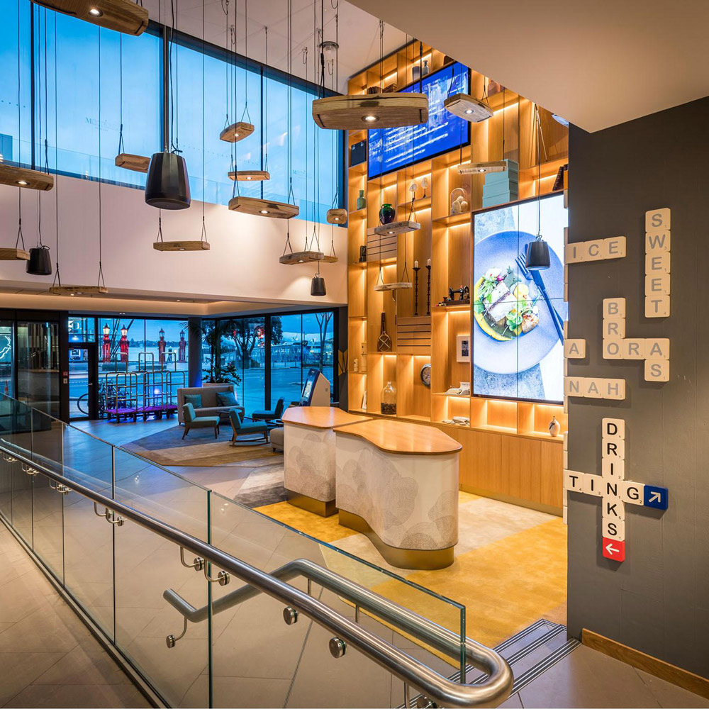 Aedas Interiors’ M Social in Auckland is shown as a vibrant double‑height hotel lobby and dining space with layered timber shelving, illuminated display walls, and sculptural pendant lighting. The image highlights a socially driven hospitality interior oriented toward waterfront views, blending contemporary design with nostalgic local references and open, interactive public spaces that encourage movement, gathering, and casual dining.