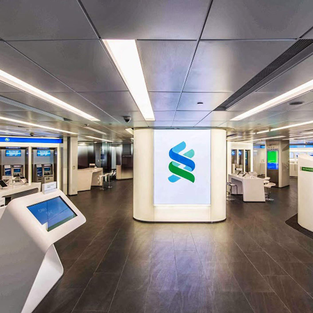 Aedas Interiors’ Standard Chartered Bank Des Voeux Road retail branch in Hong Kong is shown as a modern banking hall with self‑service kiosks, integrated digital ATMs, and a central illuminated branding column. The image highlights a secure, streamlined financial interior featuring neutral finishes, clear circulation, and technology‑enabled customer zones designed to enhance efficiency and brand visibility within a high‑traffic urban branch environment.