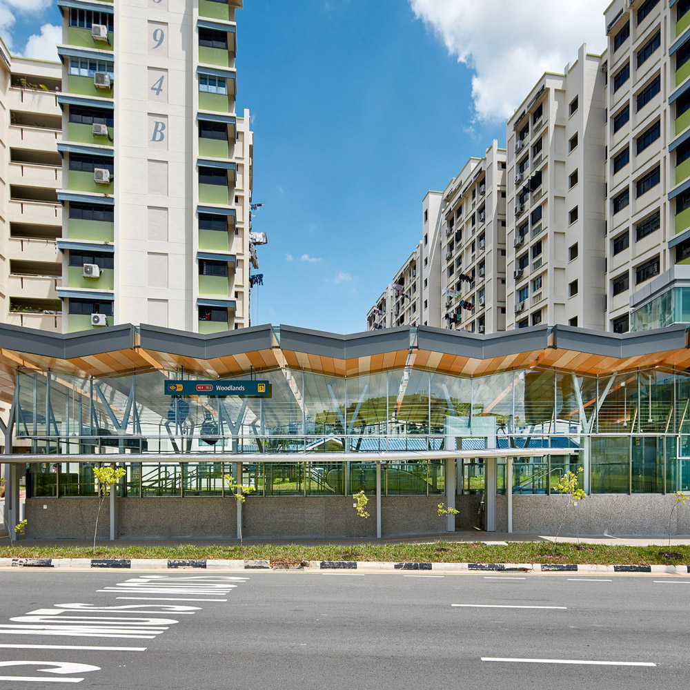 Aedas’s Woodlands Station on Singapore’s Thomson–East Coast Line is shown with a transparent glazed station box, slender steel supports, and a folded lightweight roof canopy. The exterior highlights climate‑responsive transit architecture, clear structural expression, and integration with surrounding high‑density residential buildings in a tropical urban context.