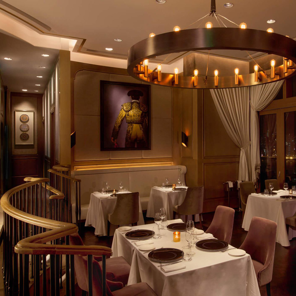 Aedas Interiors’ Zurriola in Hong Kong is shown as a contemporary fine‑dining interior with soft fabric‑upholstered seating, timber wall panelling, sheer drapery, and a circular chandelier feature. The image highlights an elegant, romantic hospitality environment defined by muted tones, refined lighting, and luxurious textures that create an intimate and serene dining atmosphere.