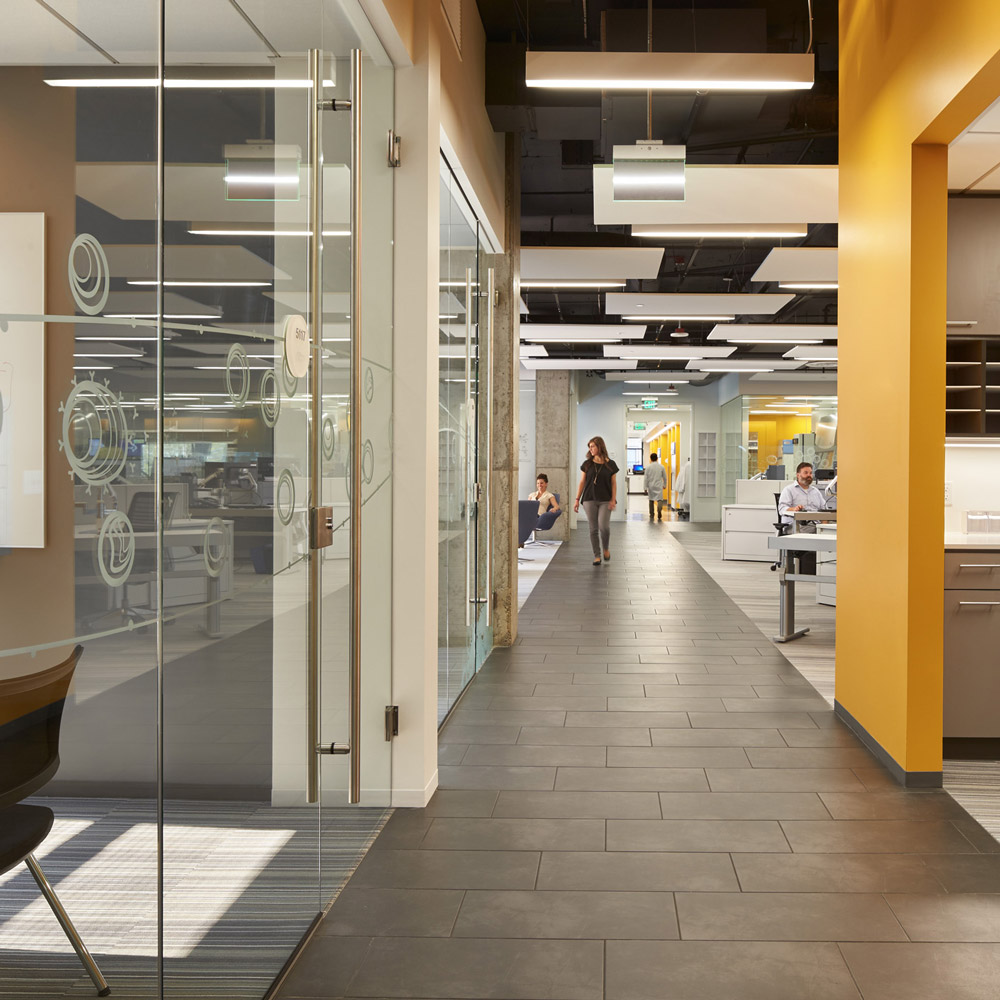 Aedas’s Celgene Seattle workplace interior is shown as an open‑plan office environment with glazed conference rooms, linear circulation spine, exposed services ceiling, and integrated workstations. The image highlights flexible research‑support architecture, transparent partition systems, and performance‑led workplace planning within a life‑sciences headquarters.