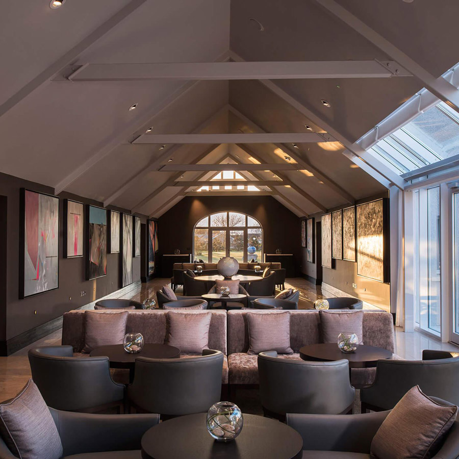 Aedas Interiors’ Twr y Felin Hotel in St Davids is shown as an elegant gallery lounge with pitched timber roof trusses, skylight glazing, polished stone flooring, and curated contemporary artworks lining dark feature walls. The image highlights a refined hospitality interior that merges historic architectural fabric with modern bespoke furnishings, creating an intimate, art‑driven sanctuary with strong visual focus and layered seating zones.