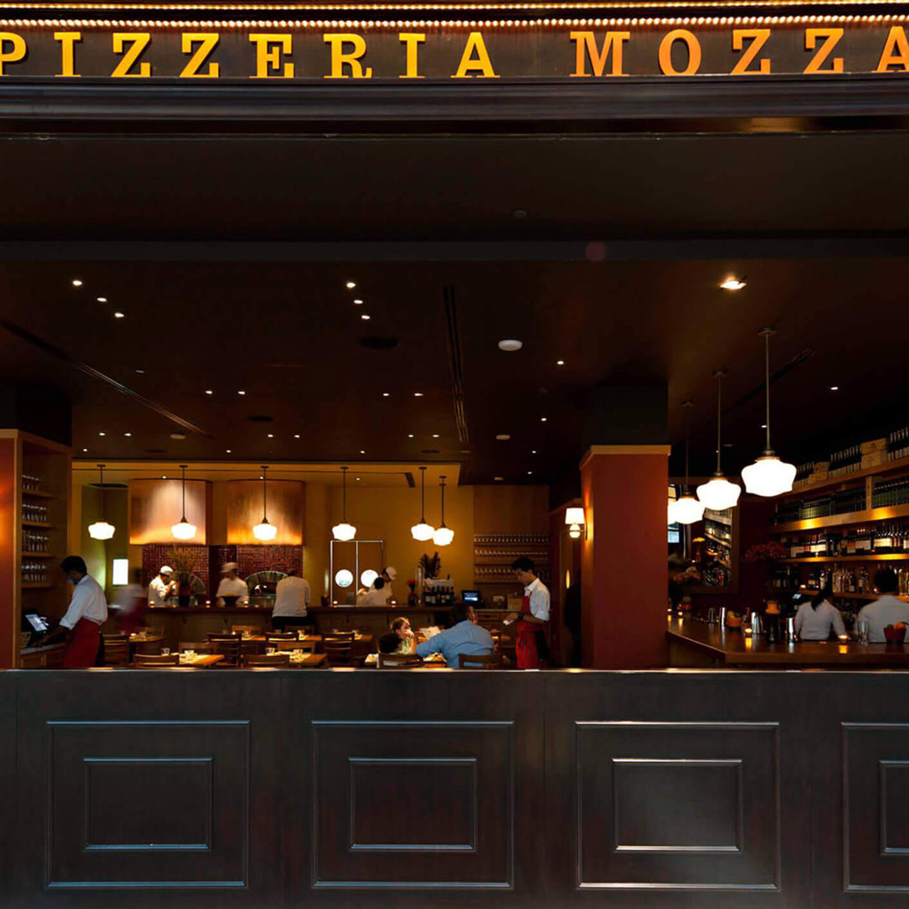 Aedas Interiors’ Pizzeria Mozza in Singapore is shown as a lively, open‑front restaurant interior with a prominent illuminated “PIZZERIA MOZZA” sign, dark timber bar counter, central white marble mozzarella bar, and warm pendant lighting. The image highlights an energetic, urban dining environment where open kitchens, wine displays, and communal seating create a vibrant hospitality experience rooted in Italian culinary tradition.