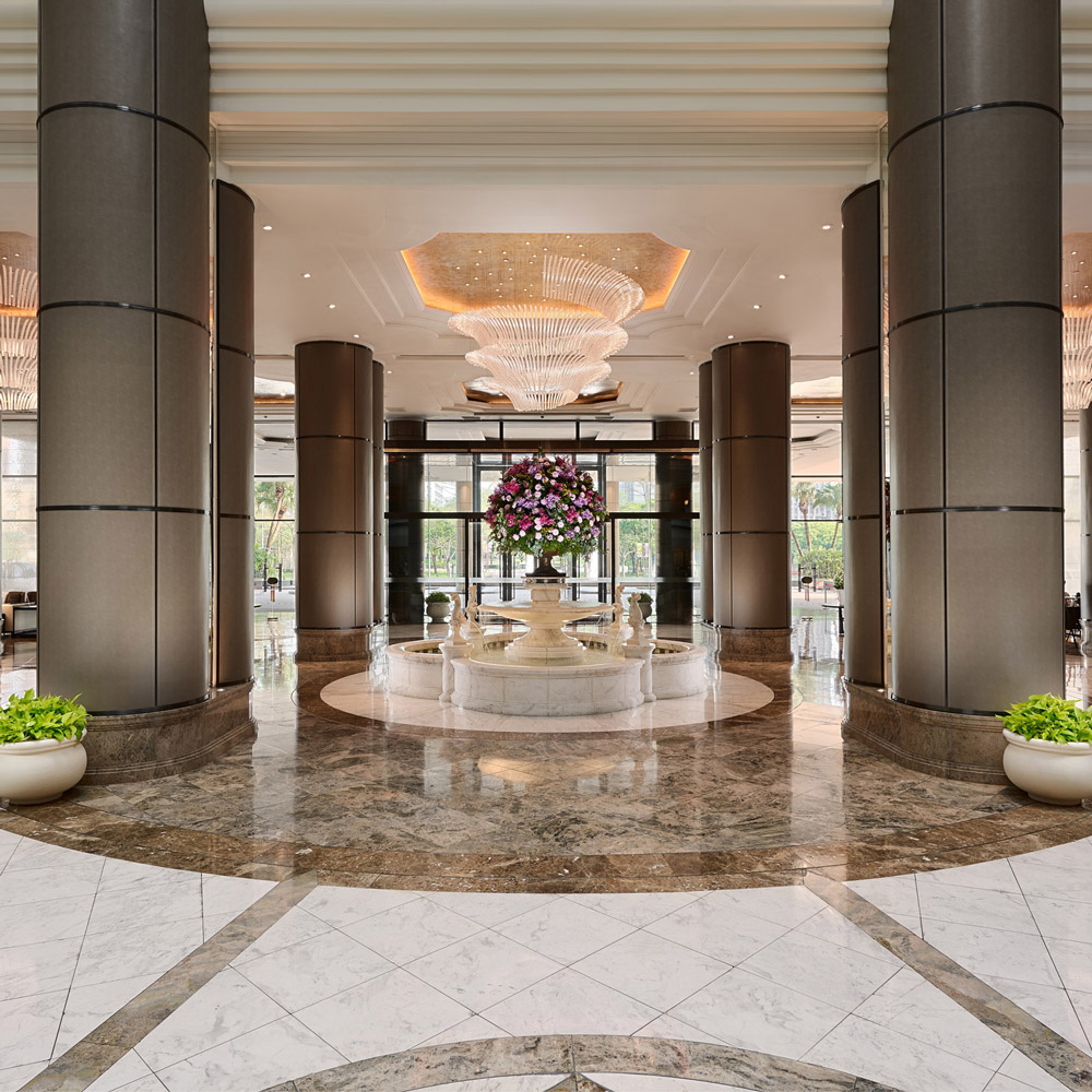 The renovated Grand Hyatt Taipei lobby is shown as a grand, symmetrical arrival hall with tall cylindrical columns, polished marble flooring, and layered coffered ceilings. Large sculptural crystal chandeliers hang above a central floral arrangement, while warm lighting and refined stone finishes create a luxurious, contemporary hospitality interior with a strong sense of scale and ceremonial arrival.