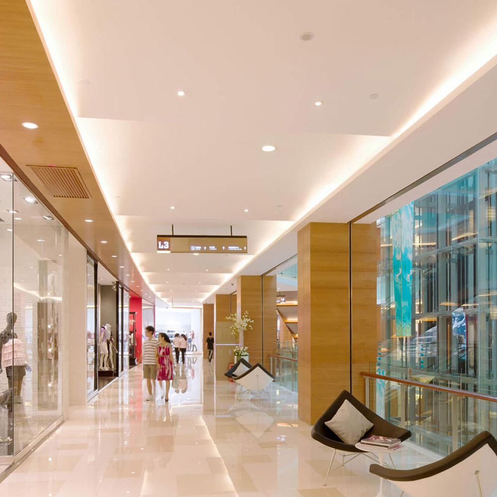 Aedas Interiors’ Renhe Spring Department Store in Chengdu is shown as a refined retail gallery with polished stone flooring, warm timber ceiling panels, full‑height glass shopfronts, and integrated lounge seating overlooking a glazed atrium. The image highlights an upscale shopping environment where natural materials, soft lighting, and generous circulation create a cosmopolitan, welcoming department‑store experience supporting international and independent brands.