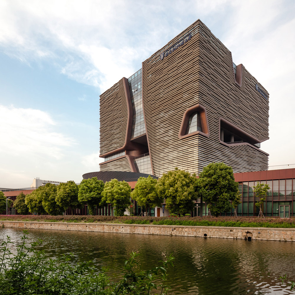 Aedas’ Jiaotong-Liverpool University Central Building in Suzhou with porous cube massing, horizontal cladding and wind‑ventilating façade voids. External view of Aedas-designed Xi’an Jiaotong‑Liverpool University Central Building in Suzhou, showing a horizontally stratified cubic volume on a landscaped plinth. The façade is wrapped in layered horizontal fins, punctured by deep sculpted voids and slots that introduce daylight and enable passive airflow through the academic building in a temperate East China climate.