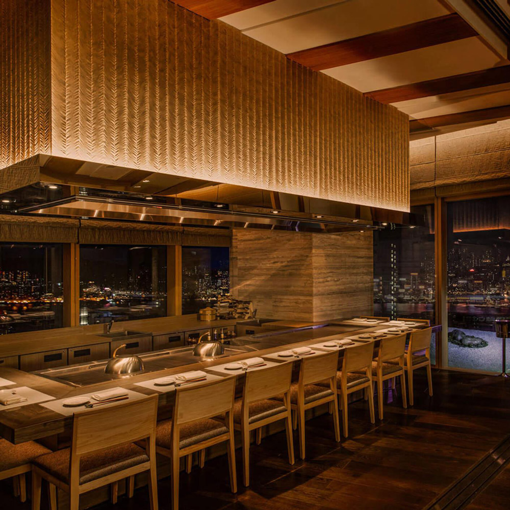 Aedas Interiors’ Kyo‑Shun in Hong Kong is shown as a refined Japanese dining interior featuring a long timber sushi counter, woven-texture feature ceiling, warm wood flooring, and minimalist seating. The image highlights a tranquil hospitality environment characterised by natural materials, subdued lighting, and craftsmanship‑driven detailing that frames the culinary experience with calm elegance.