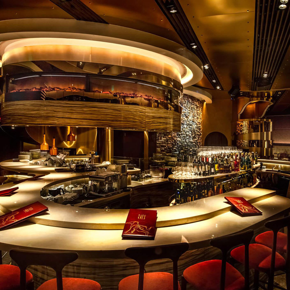 Aedas Interiors’ Tapagria in Hong Kong is shown as an energetic tapas bar interior featuring a circular marble bar counter, dark timber finishes, sculptural ceiling panels, and warm ambient lighting. The image highlights a dramatic hospitality environment designed to transition from dining to late‑night lounge, combining rich materials, open kitchen theatrics, and intimate bar seating.