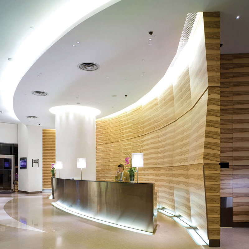 Aedas Interiors’ Novotel Century Hong Kong lobby is shown as a fluid, contemporary arrival space with a curved metallic reception counter, layered stone‑textured wall panels, and sculptural ceiling lighting. The image highlights a water‑inspired hospitality interior where soft curves, reflective surfaces, and warm illumination create a calm yet dynamic focal point within the hotel’s public areas.