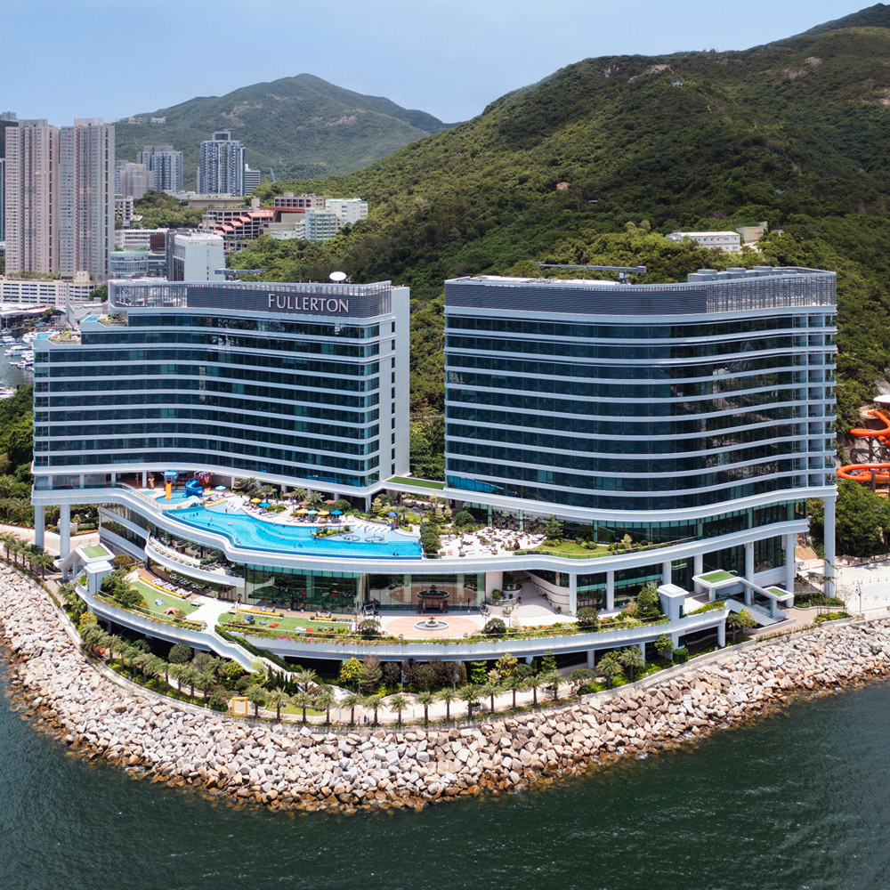 Aedas’ Fullerton Ocean Park Hotel with twin streamlined towers and cascading podium terraces along Hong Kong’s south coast. Aerial view of the Aedas‑designed Fullerton Ocean Park Hotel Hong Kong, showing two curved hotel towers with horizontal façade banding atop a stepped podium. The cascading terraces face the South China Sea, integrating outdoor pools and landscaped decks, while the complex is embedded between the rocky shoreline and the green hillside of Brick Hill.