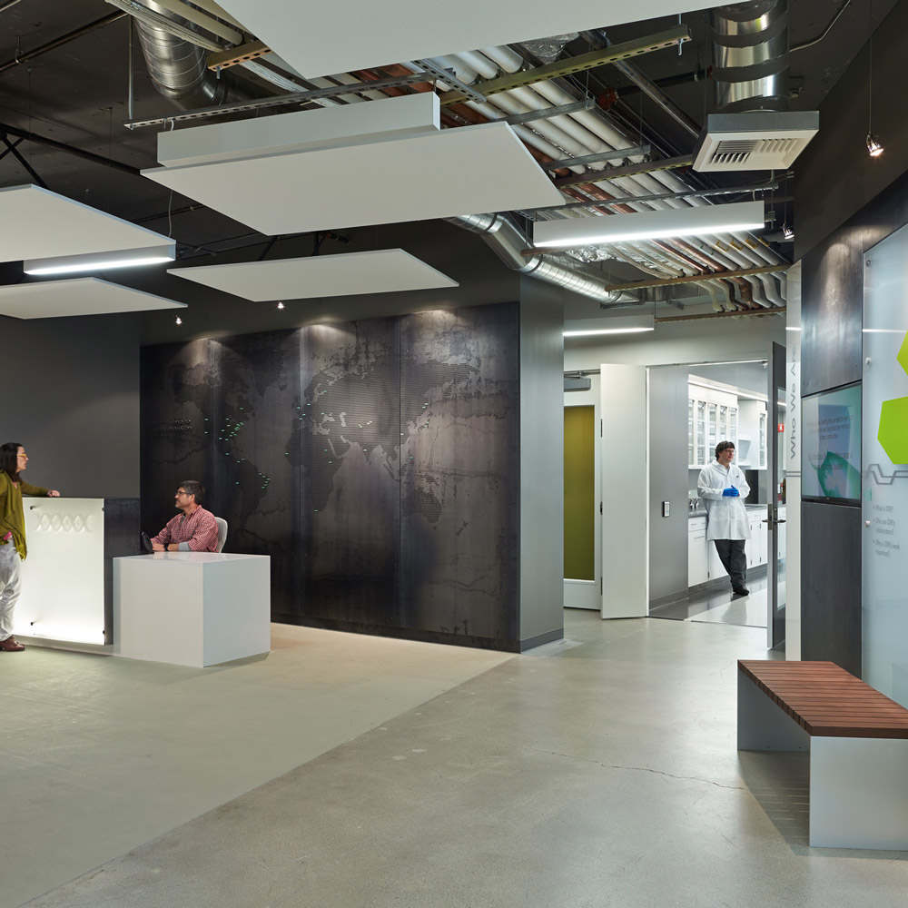 Aedas’s Infectious Disease Research Institute in Seattle is shown with an open research workplace featuring exposed concrete structure, visible mechanical services, suspended acoustic ceiling panels, integrated lighting, and flexible modular partitions. The interior highlights laboratory‑adjacent office architecture designed for collaboration within a controlled research environment.