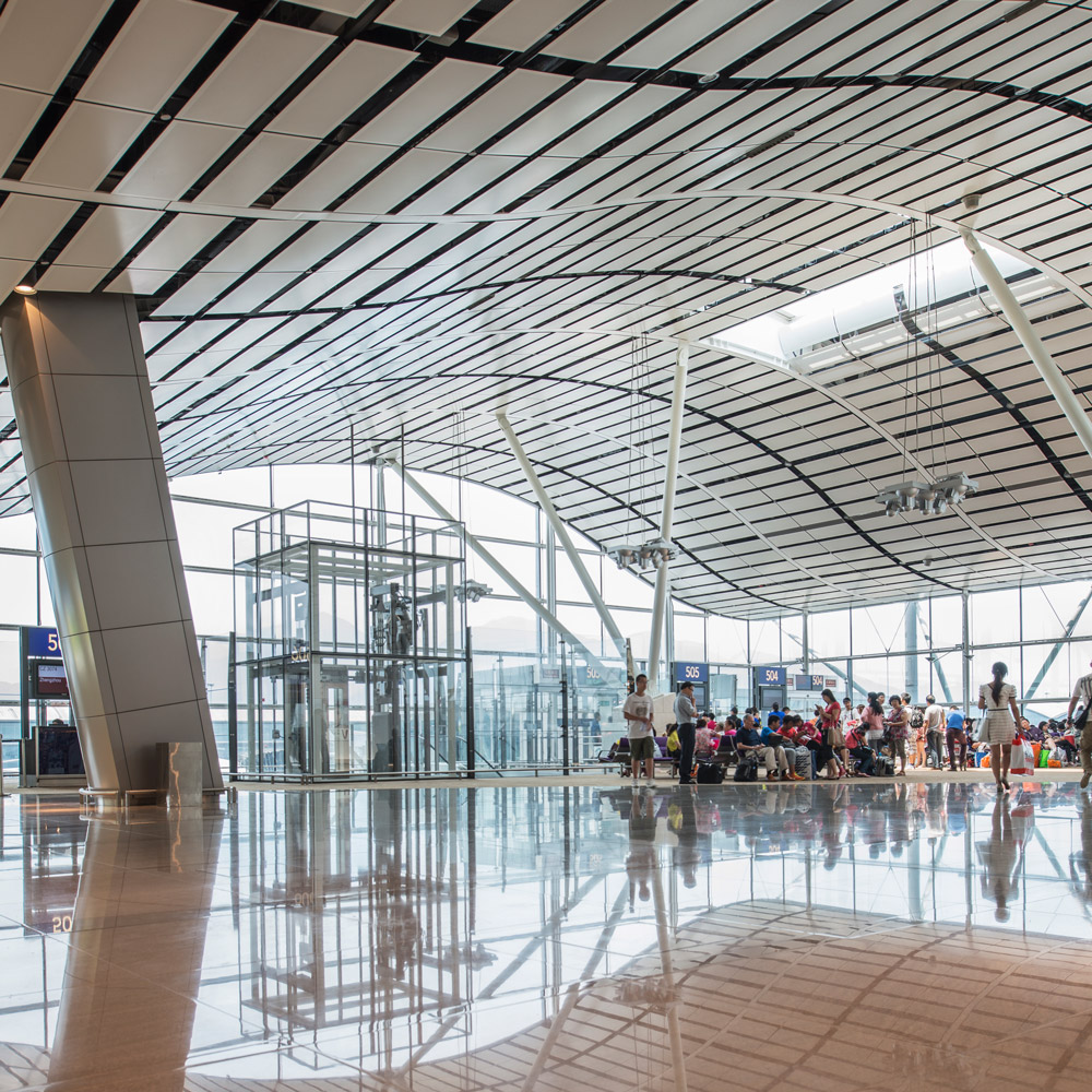 Aedas’ North Satellite Concourse at Hong Kong International Airport, showing winged roof, exposed steel columns and full‑height glazing. Interior view of Aedas-designed North Satellite Concourse at Hong Kong International Airport, featuring a curved winged roof with linear ceiling baffles, inclined steel support columns and full-height glazing. Polished stone flooring reflects natural daylight across the open passenger hall, with seating zones, vertical circulation cores and clear sightlines supporting transfer and boarding operations.