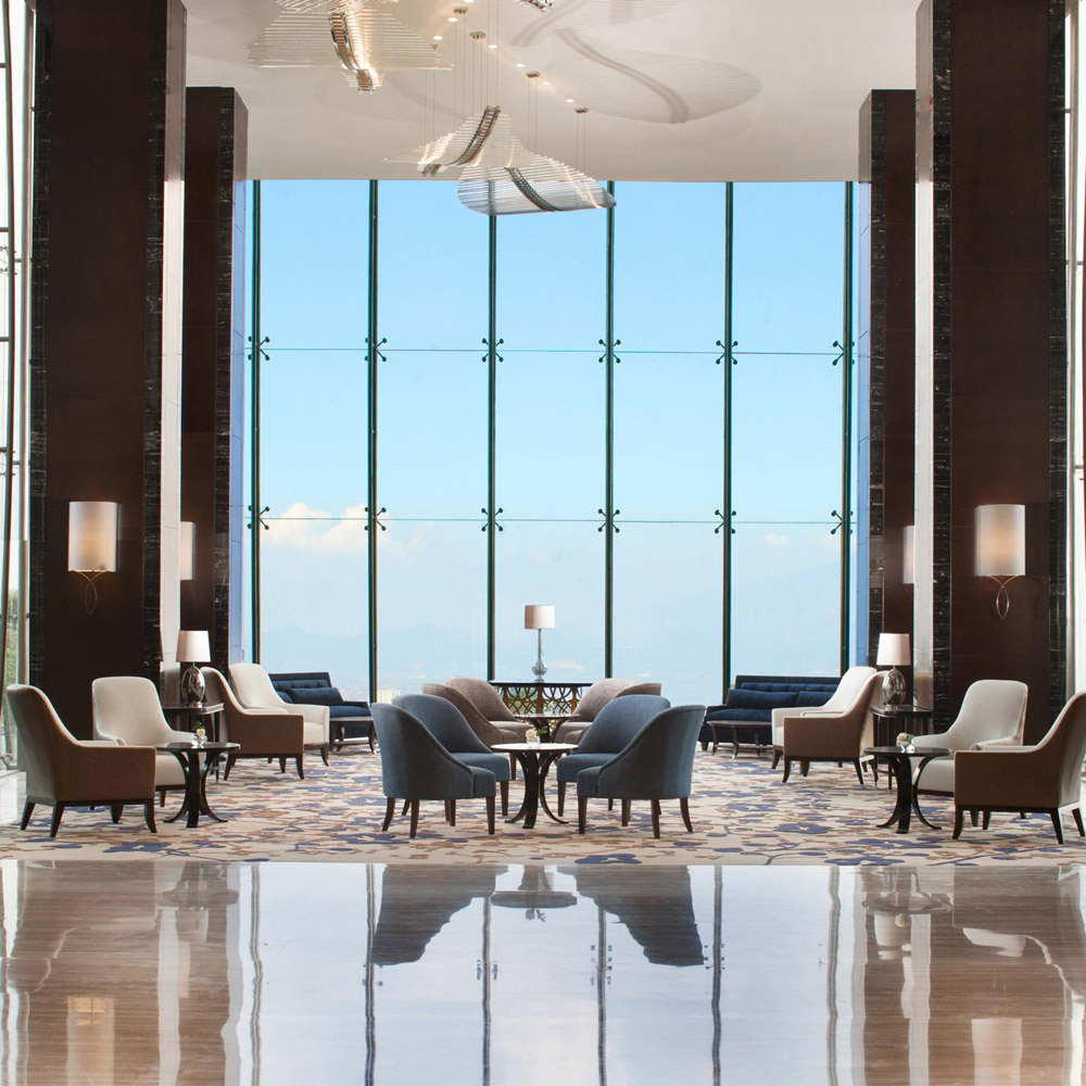 Aedas Interiors’ InterContinental Hotel Bandung is shown as a grand hotel lobby with floor‑to‑ceiling glazing framing natural views, polished stone flooring, dark timber wall panels, and sculptural ceiling art. The image highlights a serene hospitality interior that balances contemporary luxury with a restrained material palette, allowing landscape and light to define the spatial experience.