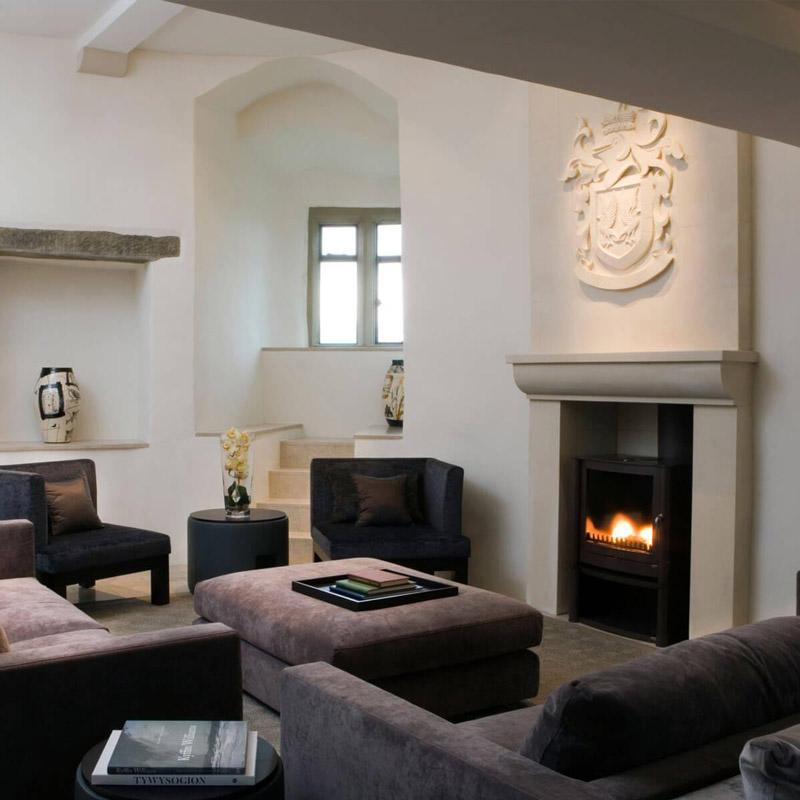 Aedas Interiors’ Roch Castle Hotel in Haverfordwest is shown as an intimate historic lounge with thick whitewashed stone walls, a sculpted stone fireplace, soft upholstered seating, and a glowing hearth. The image highlights a carefully restored medieval interior where restrained contemporary furnishings, heritage masonry, and curated artifacts create a calm, atmospheric hospitality space that foregrounds the castle’s history and sense of sanctuary.