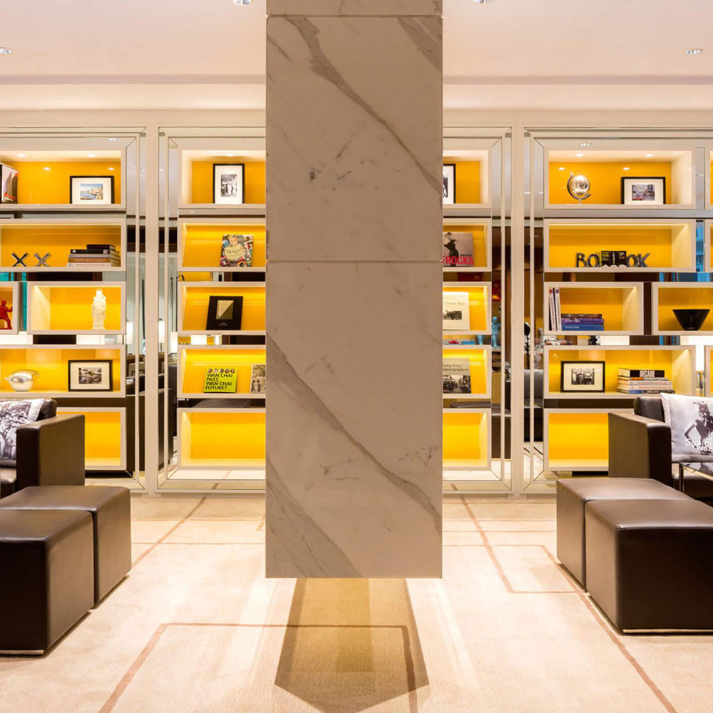 Aedas Interiors’ Hotel Indigo Hong Kong Island is shown as an intimate lounge setting with symmetrical built‑in shelving illuminated in warm yellow, marble column framing, and low leather seating. The image highlights a boutique hospitality interior that layers curated objects, graphic books, and soft lighting to express local character while maintaining a refined, residential‑style atmosphere within a dense urban hotel environment.