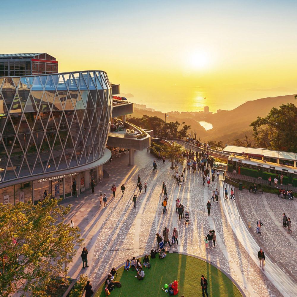 Aedas’s Peak Galleria in Hong Kong is shown as a hilltop retail destination with a faceted glazed façade, elevated pedestrian plaza, and stepped circulation. The image highlights transparent enclosure systems, public‑realm integration, and landscape‑responsive architecture positioned along Victoria Peak’s transport and viewing corridor.
