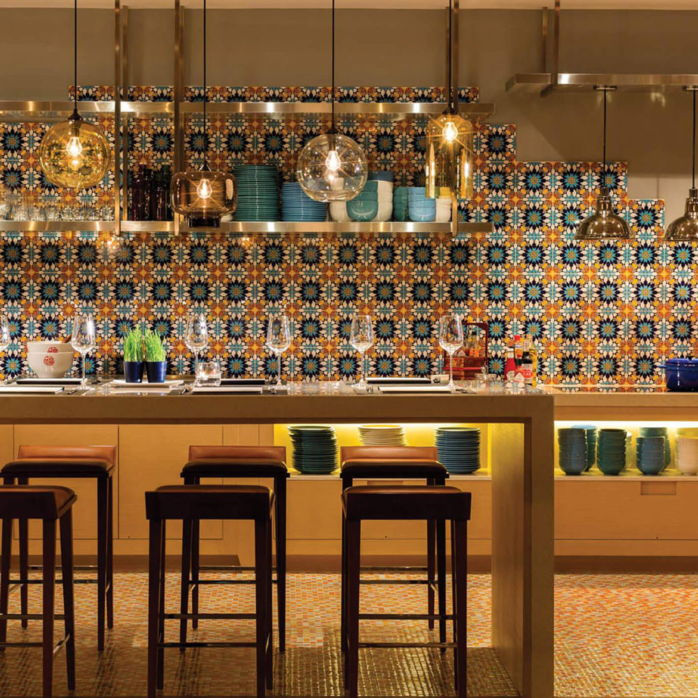 Aedas Interiors’ Café Post at Hotel Indigo Hong Kong Island is shown as a casual all‑day dining interior with an open kitchen bar, patterned ceramic tile backsplash, exposed shelving of tableware, and warm pendant lighting. The image highlights a welcoming, home‑like restaurant environment designed for social interaction, blending contemporary finishes with references to local heritage in a relaxed hospitality setting.