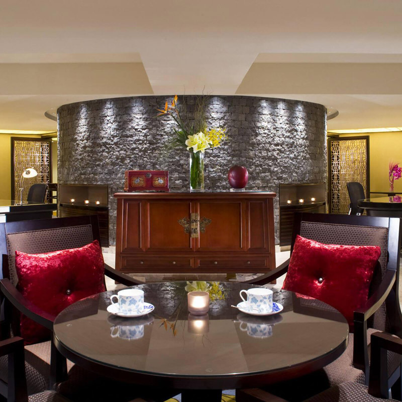 Aedas Interiors’ The Portman Ritz‑Carlton Hotel Shanghai is shown as an intimate lounge setting with curved stone feature walls, dark timber furniture, plush upholstered seating, and warm ambient lighting. The image highlights a refined hospitality interior blending Eastern and Western influences through layered materials, controlled symmetry, and elegant detailing designed to support comfort and privacy within a luxury hotel environment.