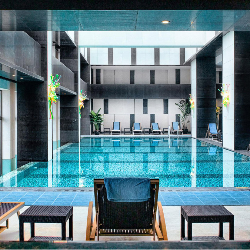 Aedas Interiors’ Ximei Business Hotel in Shijiazhuang is shown with a tranquil indoor rooftop infinity pool featuring gradient mosaic tiling, dark stone‑clad columns, and lounge chairs arranged along the pool edge. The image highlights a contemporary hospitality leisure space combining reflective water surfaces, clean architectural lines, and controlled natural light to create a refined business‑hotel retreat overlooking the urban skyline.