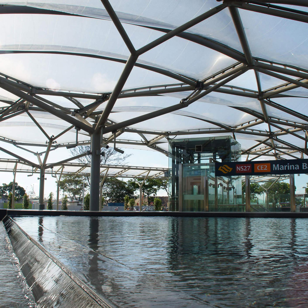 Aedas’ Marina Bay MRT Station with ETFE skylight canopy, reflective pool and glazed entrance set within parkland. Street‑level view of the Aedas‑designed Marina Bay MRT Station in Singapore, showing a lightweight steel structure supporting a translucent ETFE canopy. The canopy spans above a reflective pool and glass lift enclosure, allowing daylight penetration while maintaining visual openness to the surrounding park landscape in a tropical urban environment.