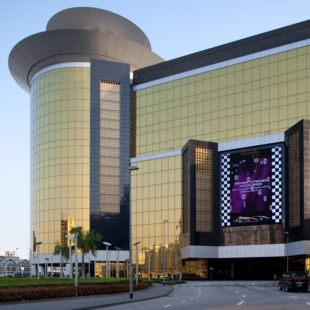 Aedas’ Sands® Macao casino resort with gold‑toned glazed façades, cylindrical hotel tower and podium entry. Street‑level view of the Aedas‑designed Sands® Macao showing a Las Vegas‑style casino podium clad in gold‑tinted curtain wall glazing, articulated by vertical black stone elements and a cylindrical tower volume above. A recessed vehicular drop‑off, landscaped forecourt and broad roadway frame the building within Macau’s dense urban resort context.